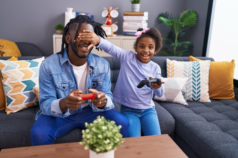 Father and daughter playing video game sitting on sofa at home | WilkComm Reno